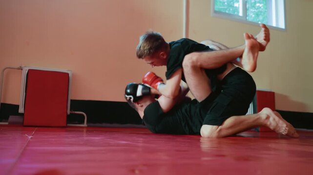 Boxers engaged in grappling exchange on red mat during sparring training, one fighter locked in guard position attempting submission while opponent maintains control, showing intensity, stamina