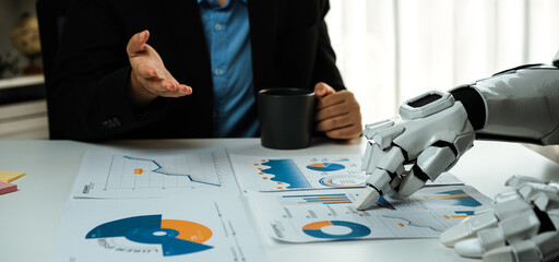 A human and a robotic arm engage in a business meeting, analyzing data charts on a desk, showcasing...
