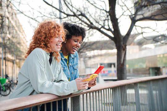 Two cheerful young women enjoying their time together as they engage with their smartphones, sharing laughter and joy while using mobile applications in a lively atmosphere. - Powered by Adobe