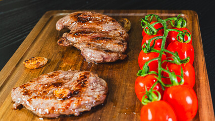 A whole, juicy grilled steak rests on a wooden board, accompanied by roasted cherry tomatoes on the vine and whole fried garlic cloves. Rustic and appetizing culinary scene.