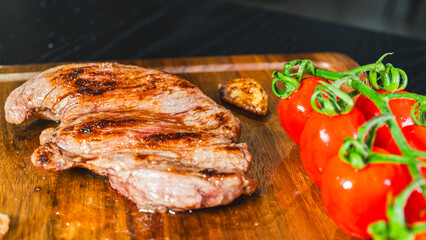 A whole, juicy grilled steak rests on a wooden board, accompanied by roasted cherry tomatoes on the vine and whole fried garlic cloves. Rustic and appetizing culinary scene.