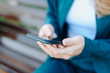 Obraz premium young woman holds smartphone in hands, texting, watching content, sitting on bench in park, close-up view, no face, copy space