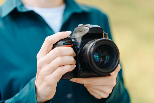black dslr camera in hands, young caucasian woman photographer in green shirt at work outdoors in sunny summer day