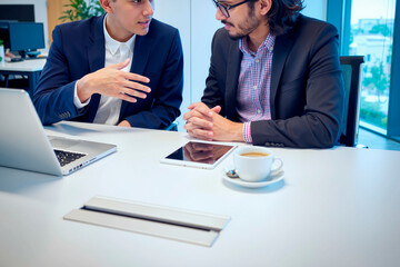 Two young adult men, one Caucasian and one possibly Hispanic, sitting at desk discussing business strategy, using laptop and digital tablet, coffee cup on table, office setting