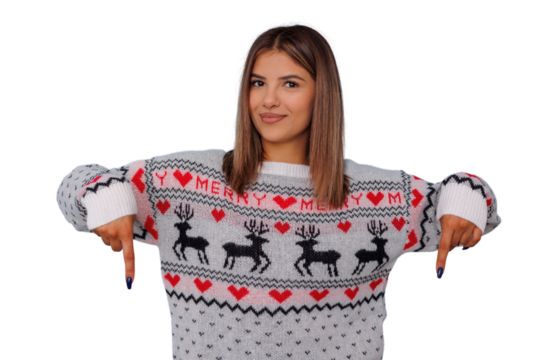 Young woman wearing festive knit sweater with reindeer and hearts, pointing both hands down, transparent background