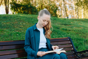 Obraz premium young beautiful redhead caucasian woman holds thick book in hands, reading book, sitting on bench in park, sunny summer day
