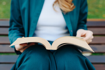 young woman holds thick book in hands, reading book, sitting on bench in park, close-up view, no...