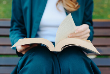 young woman holds thick book in hands, reading book, sitting on bench in park, close-up view, no...