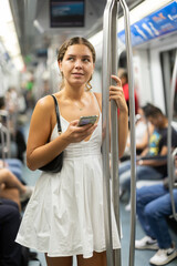 Young woman in a white dress is riding the subway, holding a mobile phone and looking around. Using of public transport