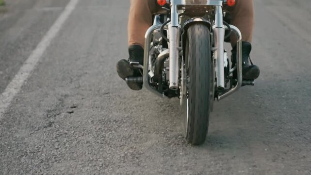 Lower section view of motorcycle rider cruising on asphalt road showing close-up of front wheel, metal frame and biker boots pressing pedals while vehicle moves steadily along countryside
