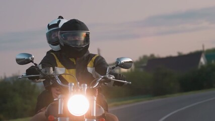 Close up of two motorbike riders wearing helmets and jackets navigating bend on empty road during evening ride through countryside, passing houses under soft twilight sky with warm headlight glow