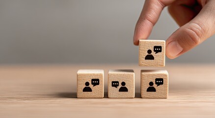 A hand places a small wooden block, featuring a symbol of two people conversing, on top of a stack of similar blocks