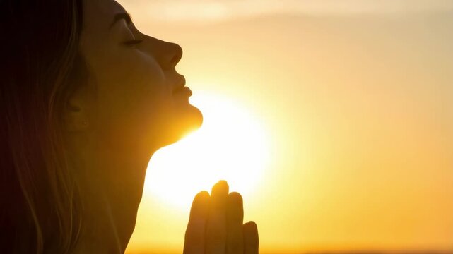 Woman in thoughtful contemplation, hands together, silhouetted by the golden glow of a rising or setting sun. Peaceful, spiritual moment.