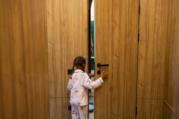 A young girl in pajamas stands at a partially ajar door, reaching out to open it. Her expression conveys curiosity and excitement about what lies beyond the threshold.