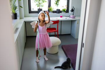 A young girl dressed in a pink fairy costume plays joyfully with a wand while her father watches and smiles. A cat observes the fun nearby, creating a heartwarming family atmosphere.