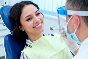 Young adult Caucasian woman sitting in dental chair smiling while male dentist wearing face shield and gloves examining teeth with dental mirror during checkup