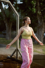 A woman stands in a park, soaking up the sun while wearing a yellow top and pink pants. She smiles and spreads her arms, enjoying a joyful moment in nature on a warm day.