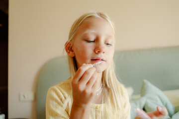 A young girl with long blonde hair sits on a bed, carefully applying lip gloss. Soft sunlight filters into the room, creating a warm atmosphere as she enjoys her beauty routine.