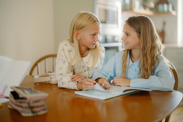 Two young girls sit closely at a wooden table, engaged in studying and sharing ideas. They work on notebooks, showcasing friendship and teamwork in a warm, sunny indoor setting.