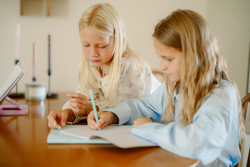 Two young girls are focused on their studies at a wooden table in a well-lit room. They are sharing ideas and working on homework together, fostering a collaborative learning environment.