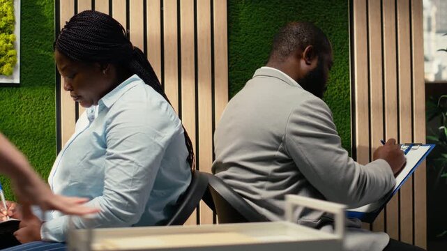 Confident job applicants filling in documents with their details, sitting in the hallway back to back before hiring interview. Reviewing CVs and preparing for employment questions with HR. Camera B.