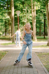 A girl is skating joyfully on a paved path in a lush green park. She wears blue overalls and smiles as she glides past trees on a sunny day. Another skater is visible in the background.