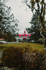 White institutional building with a prominent red roof and central tower, framed by dark green pine trees and a grassy lawn.