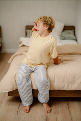 Fototapeta premium A young boy with curly hair sits on a bed wearing comfortable clothing. The bedroom has a warm atmosphere with soft lighting, creating a relaxing environment.