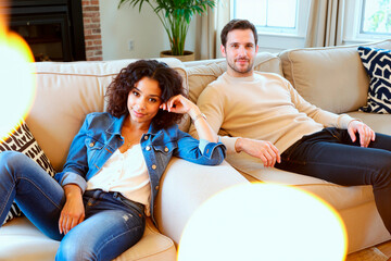 Caucasian man and Black woman relaxing on sofa in living room, both sitting comfortably and looking toward camera, casual posture suggesting leisure and connection at home
