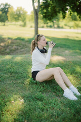 Obraz premium A young woman sits on the grass in a park, sipping from a bottle. She wears casual sports attire and headphones, enjoying a sunny day surrounded by greenery.