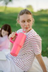 A young woman leans back while sipping a bright pink drink from a large cup, enjoying a picnic on a sunny day in a park with green grass. Another person is seated nearby.