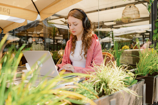 A young woman wearing headphones sits at a laptop in a bright outdoor cafe. She is focused on her work, with plants and flowers creating a vibrant atmosphere around her.