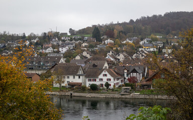 autumnal village view of quaint traditional swiss or german homes on a hillside above a tranquil river or lake under a cloudy sky, framed by colorful fall foliage
