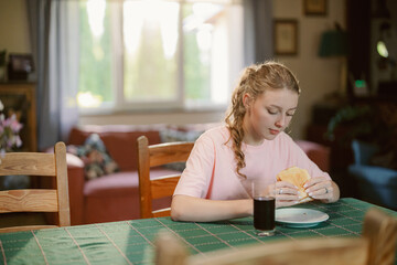 A girl sits at a table in her cozy home, savoring a sandwich. Sunlight filters through the window,...