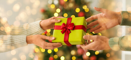 Hands of young man and woman with gift box near Christmas tree at home, closeup