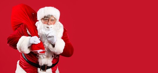 Santa Claus with bag full of Christmas gifts showing silence gesture on red background with space for text
