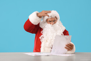 Shocked Santa Claus reading letter at table on blue background
