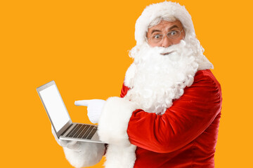 Shocked Santa Claus pointing at laptop on yellow background