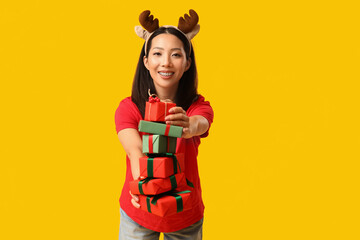 Young Asian woman in reindeer horns with Christmas gift boxes on yellow background