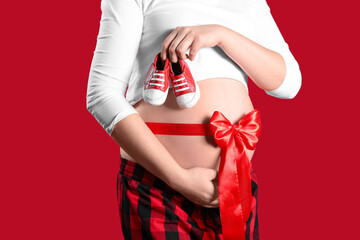 Beautiful young pregnant woman with baby shoes and ribbon around her waist on red background