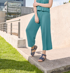 Minimalist urban summer look with woman in teal wide-leg pants and matching top, wearing blue strappy leather sandals, walking on concrete ledge beside metal railing and green grass near buildings.