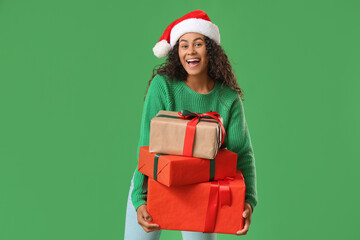 Beautiful young happy African-American woman in Santa hat with Christmas gift boxes on green background