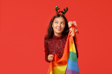 Young Asian woman in reindeer horns with LGBT flag and Christmas gift on red background