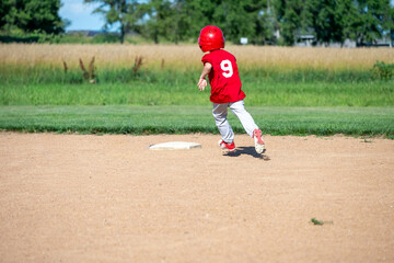 Young boy in red jersey runs towards first base during a sunny baseball game in a rural field