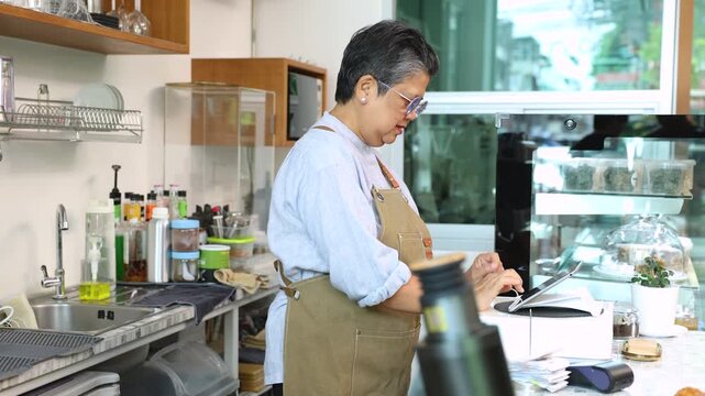 Senior entrepreneur cafe owner barista hold POS machine to customer, Asian female in apron welcome warmly in counter, croissants and glassware background, retired elderly woman start small business