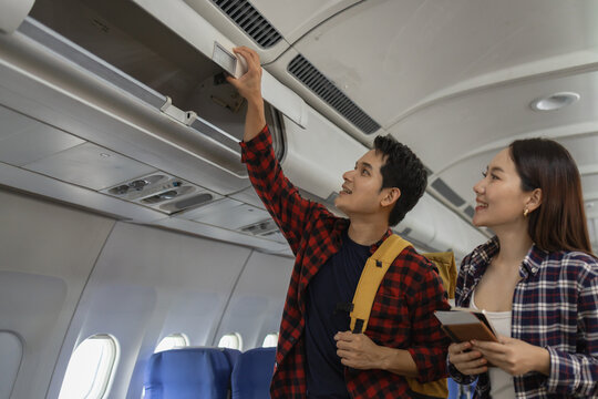 Asian couple preparing for travel on an airplane. man is putting his item into the overhead compartment while woman smiles and holds passports and boarding passes. Both look excited and ready to fly
