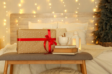 Beautiful Christmas gift, books, lantern and cup of cocoa drink on bench near comfortable bed