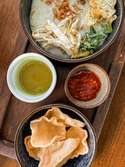 Traditional Indonesian chicken porridge or Bubur Ayam served with crackers, chili sauce, green sauce, and toppings on a wooden tray over rustic wooden table.