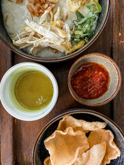 Traditional Indonesian chicken porridge or Bubur Ayam served with crackers, chili sauce, green sauce, and toppings on a wooden tray over rustic wooden table.