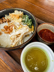 Traditional Indonesian chicken porridge or Bubur Ayam served with crackers, chili sauce, green sauce, and toppings on a wooden tray over rustic wooden table.